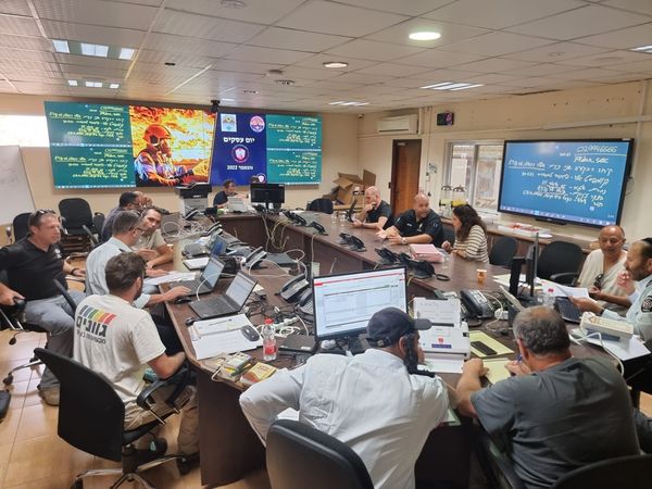 בעלי עסקים יושבים ליד אנשי כיבוי והצלה מסביב לשולחן עם מחשבים ודפים, על הקיר תלוי מסך עם הנחיות