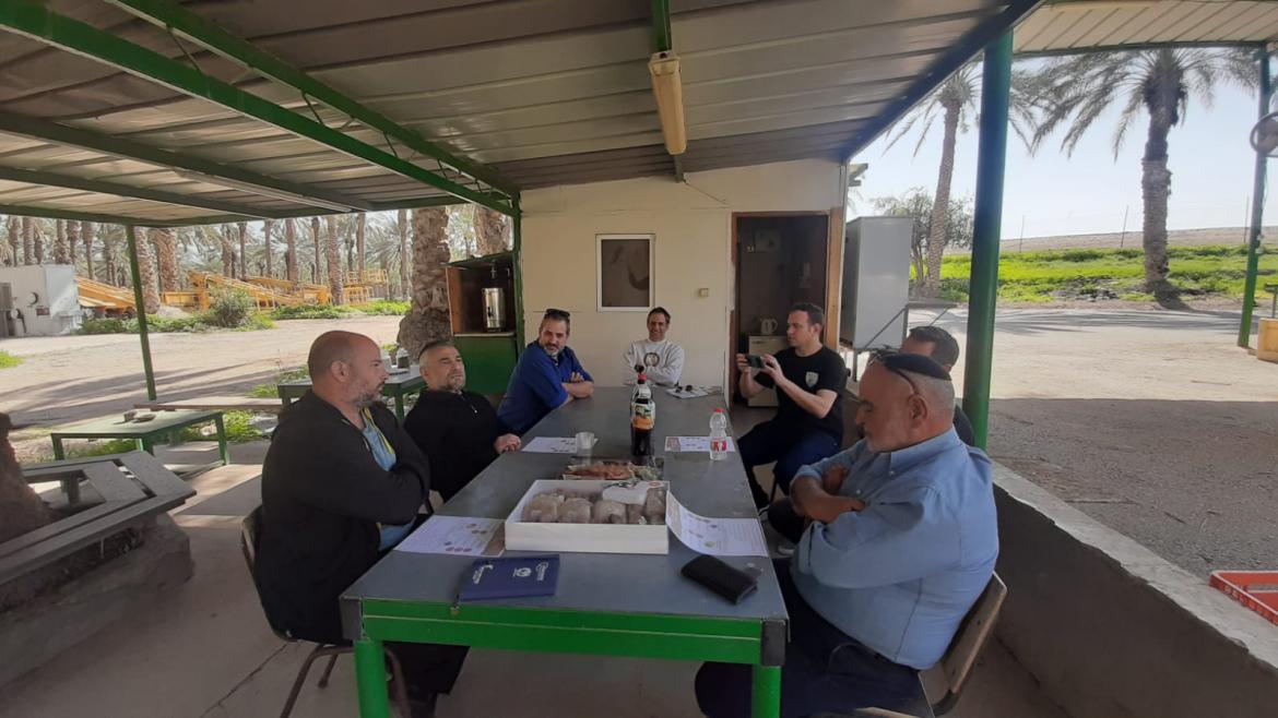 סגן השר ואנשיו יושבים ליד השולחן עם כיבוד בסככה כשברקע מטע תמרים 