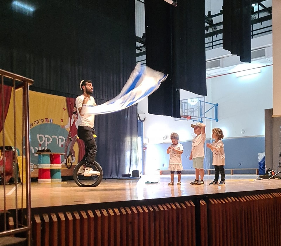 עובד קרקס על חד אופן על הבמה עם 3 ילדי גנים 