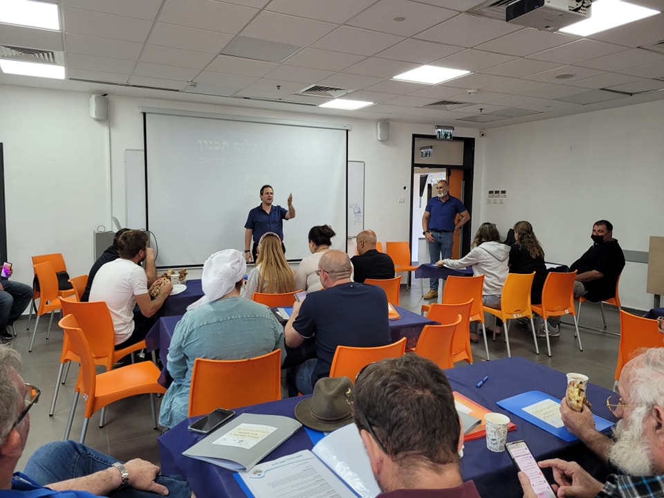 מוטי אבנר נואם בפני נציגי הישובים, ראש המועצה עומד בצד כשכולם נמצאים באולם המתנ"ס