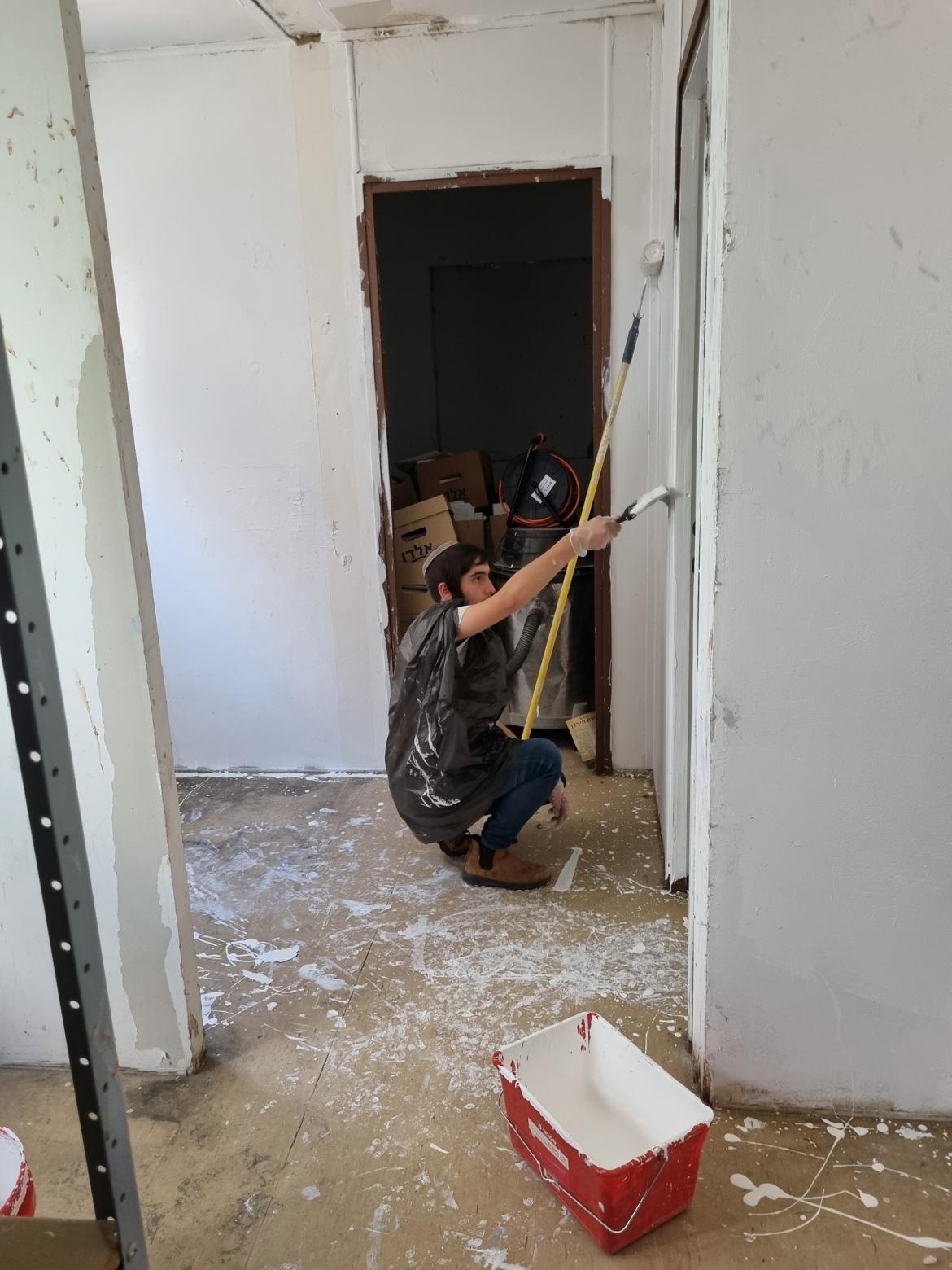 ילד צובע קיר בצבע לבן עם מכחול , כל הרצפה עם שאריות של צבע וקופסת צבע על הרצפה