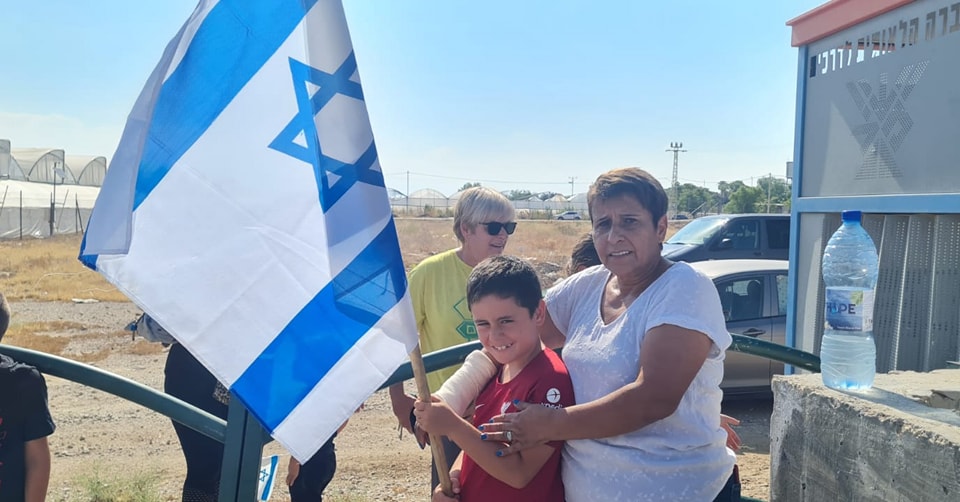 אישה עם ילד שאוחז בדגל ישראל עומדים ליד תחנת אוטובוס, ברקע עוד אנשים ומכוניות חונות
