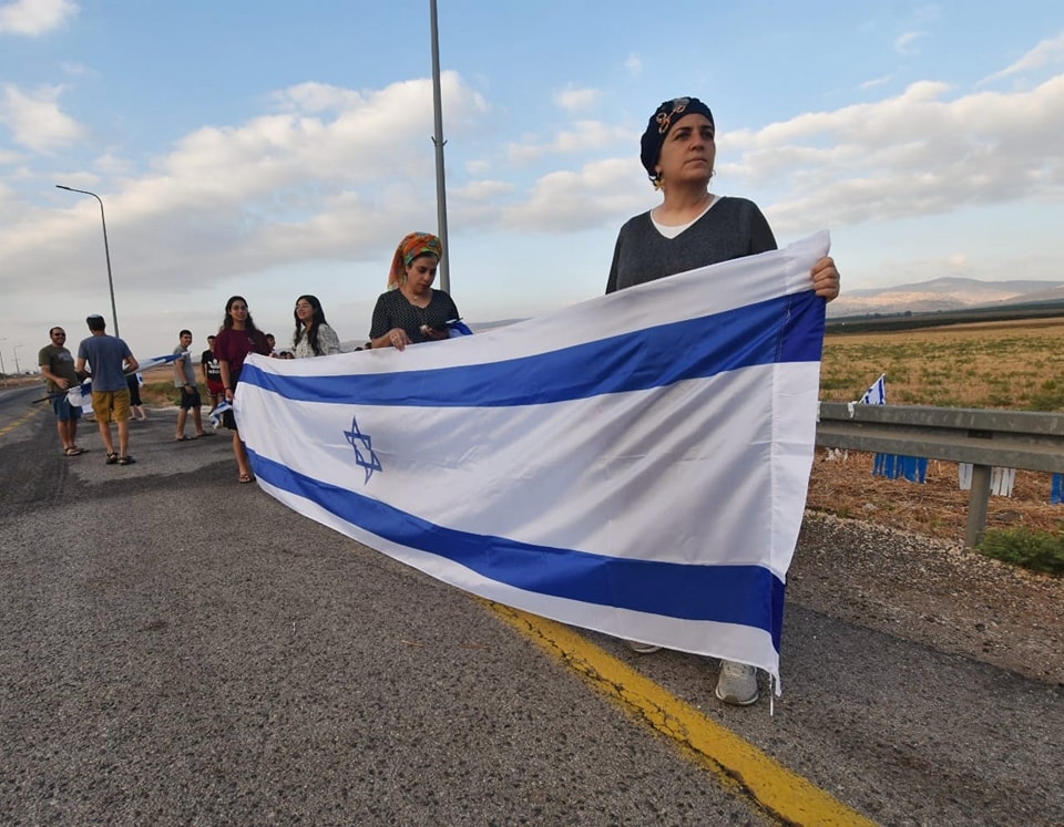 3 נשים אוחזות בדגל ישראל גדול לרוחב כשהן עומדות לצידי הכביש , ברקע עוד אנשים