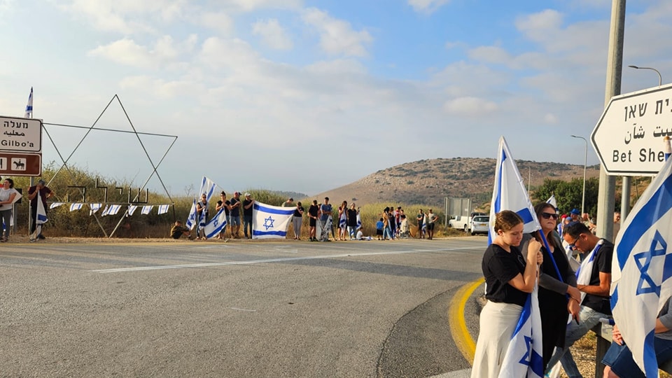אנשים עומדים בשני צידי הכביש עם דגלים, ברקע שלט "בית שאן"