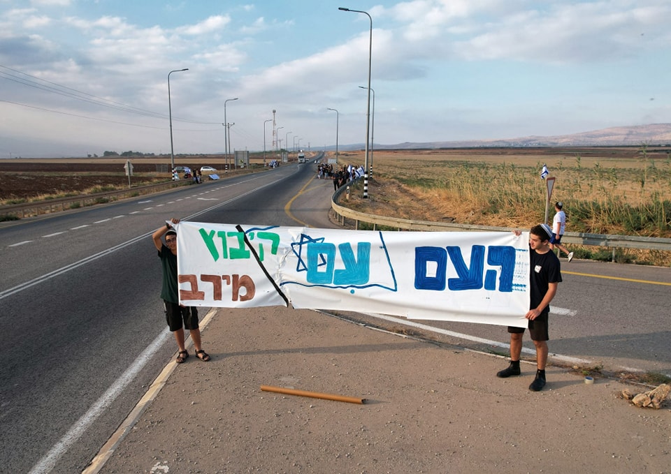 2 ילדים עומדים לצידי כביש ואוחזים בשלט: "העם עם קיבוץ מירב"