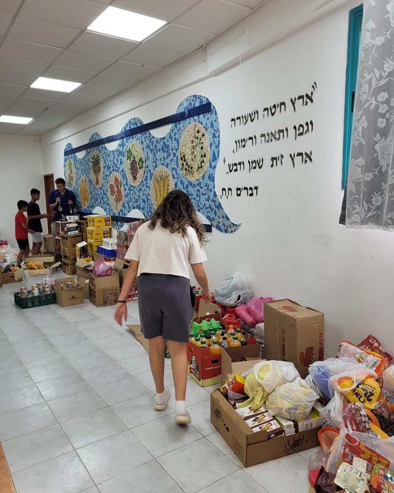 על הרצפה בתוך המועצדון קופסאות עם מצרכי מזון לחלוקה, בחורה עוברת בין הקופסאות 