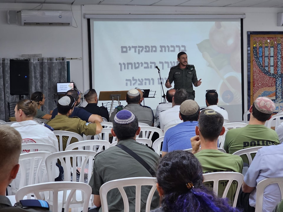 מפקד מג"ב נואם בפני המתנדבים שיובים וברקע מסך עם כתובית: ברכות מפקדים מערך הבטחון, חירום והצלה