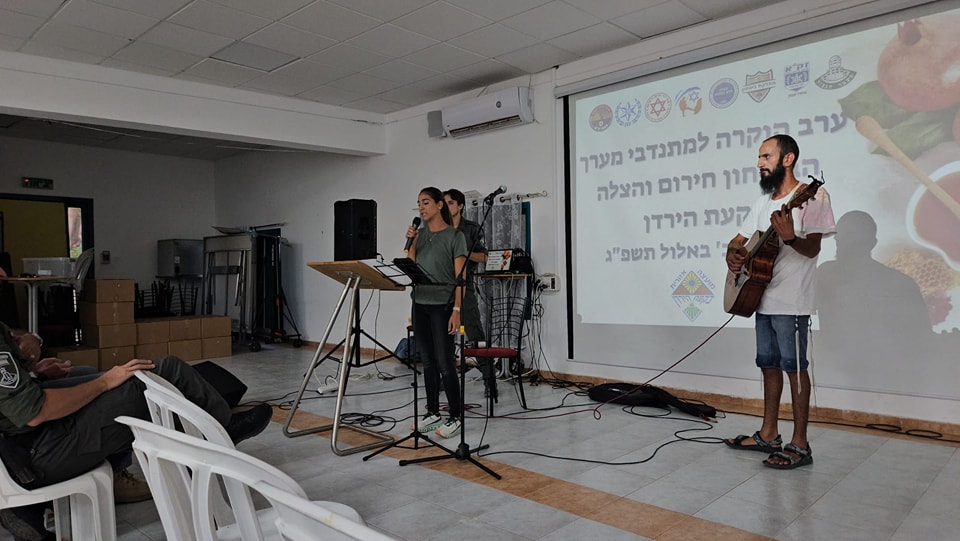 קבטית המועצה נואמת , לידה בחור מנגן בגיטרה וברקע מסך עם הכתובית: ערב הוקרה למתנדבי מערך הבטחון, חירום והצלה בקעת הירדן